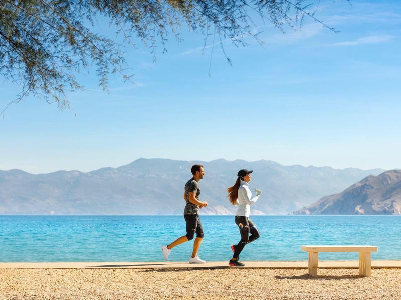 Zwei Personen joggen am Sandstrand mit Meer und Bergen im Hintergrund unter blauem Himmel.