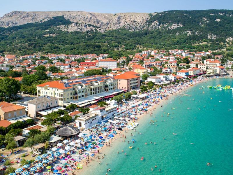 Küstenstadt mit Strand, türkisfarbenem Wasser, bunten Sonnenschirmen und Bergen im Hintergrund.
