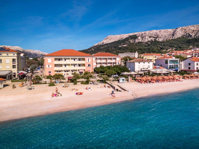 Strand mit türkisfarbenem Wasser, Sand, Liegestühlen, Gebäuden und Bergen im Hintergrund.
