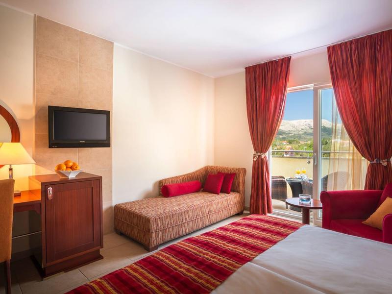 Helles Hotelzimmer mit Bett, rot gestreifter Bettdecke, Sofa, Schreibtisch, TV und Balkon mit Aussicht.