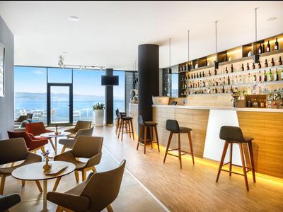 Modern hotel bar area with wooden counter, seating, and large window with sea view.