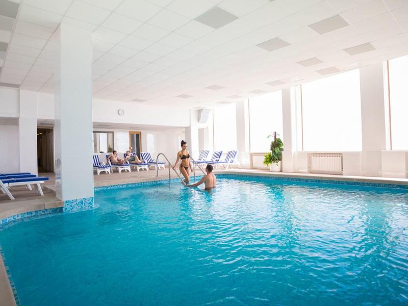 Indoor pool with relaxing guests and lounge chairs in bright room with large windows.