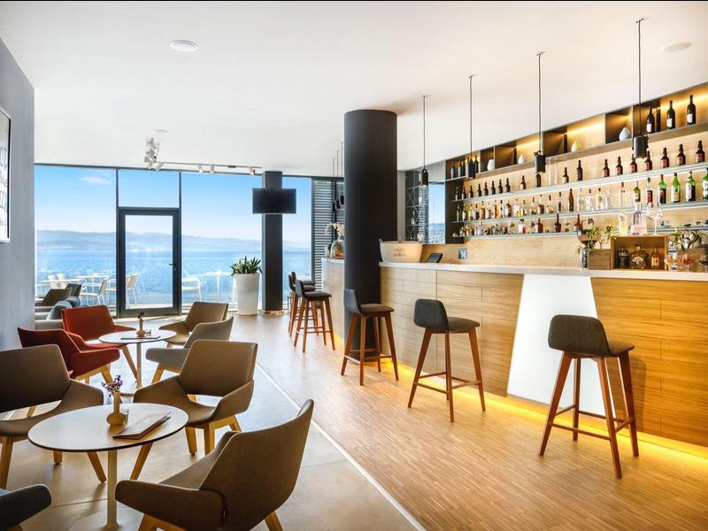 Modern hotel bar area with wooden counter, seating, and large window with sea view.