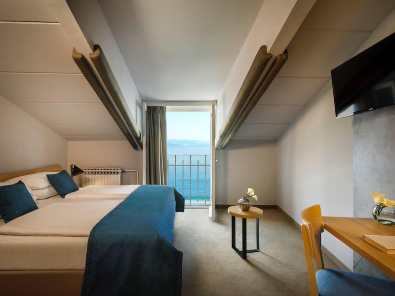 Modern hotel room with double bed, balcony, and sea view under natural light.