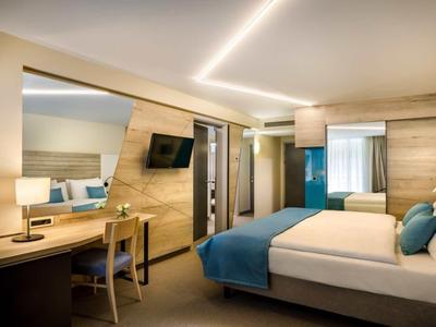 Modern hotel room with double bed, desk, chair, illuminated mirror, and wall-mounted TV.