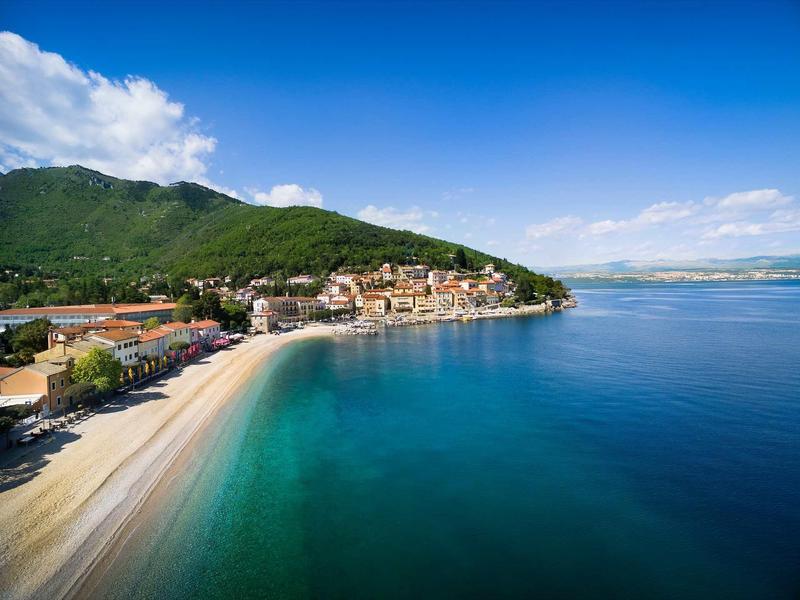 Beach with clear water and coastal town by forested hills under clear sky