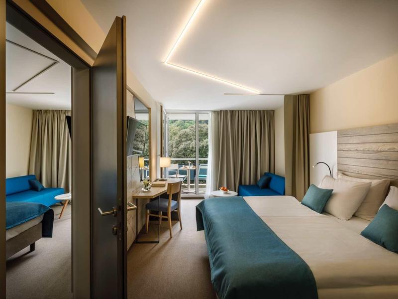 Modern hotel room with two beds, desk, and balcony view of natural surroundings.