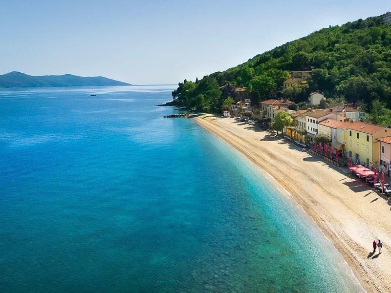 View of a long sandy beach next to clear blue water with a wooded hill and buildings.