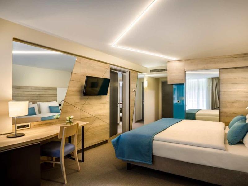Modern hotel room with double bed, desk, chair, illuminated mirror, and wall-mounted TV.