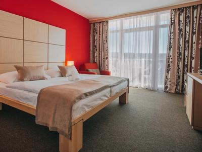 Helles Hotelzimmer mit Doppelbett vor roter Wand und Balkon mit Vorhängen