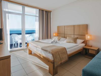 Helles Hotelzimmer mit Doppelbett, Fernseher, Sofa und Balkon mit Meerblick.