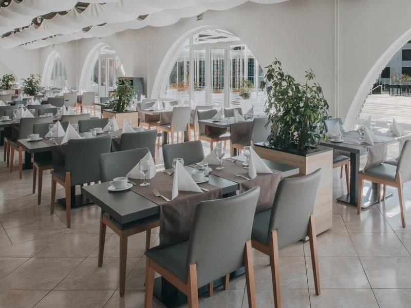 Bright restaurant with gray chairs, wooden tables, and large windows overlooking a building.