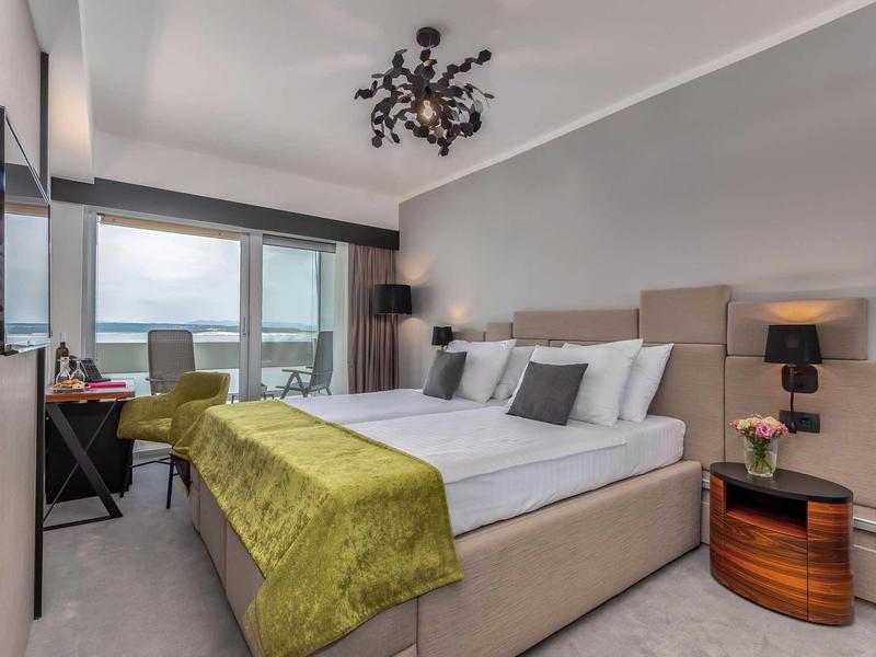 Modern hotel room with large bed, green throw, and balcony overlooking the sea.