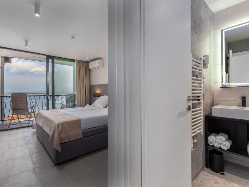 Modern hotel room with double bed, balcony overlooking the sea, and stylish bathroom.