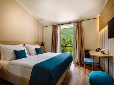 Brightly furnished hotel room with double bed, desk, and view of green mountain landscape.