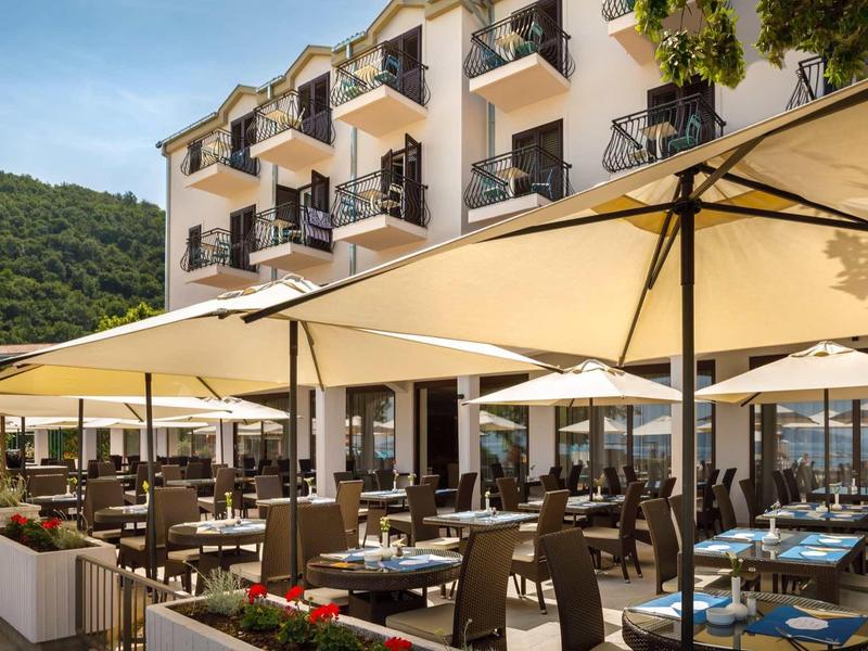Outdoor area of a hotel with many tables, chairs, and large umbrellas in front of a multi-story building.