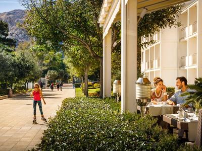 Lidé sedí venku v kavárně poblíž hotelu, obklopeni stromy a horami.