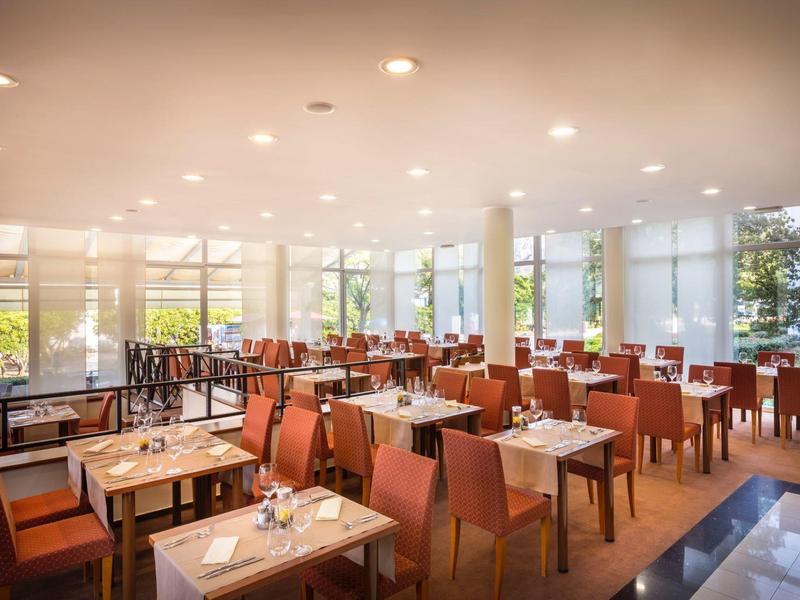 Heller, moderner Speisesaal mit Holztischen und roten Stühlen, großen Fenstern und Deckenleuchten.