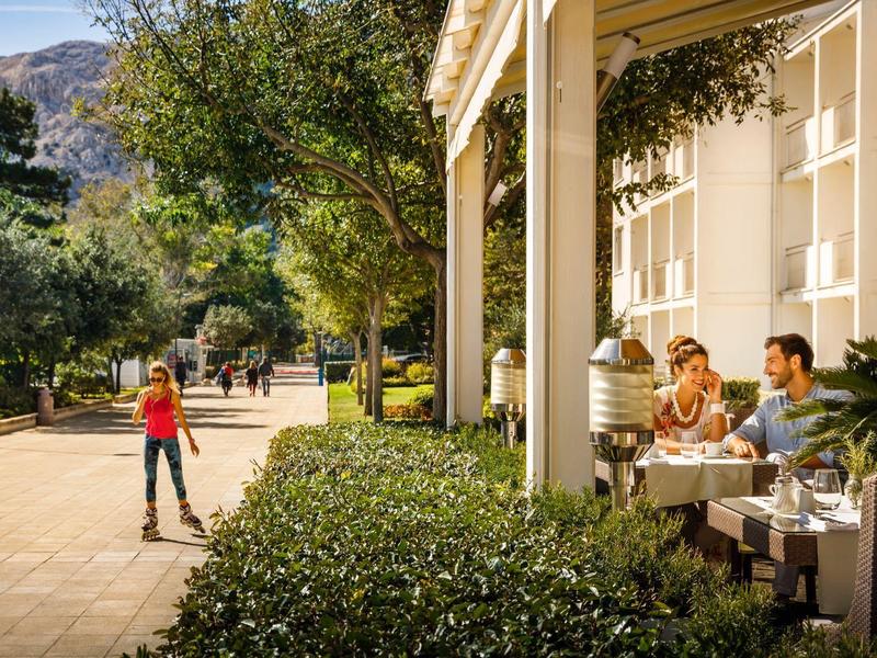 Mann mit Hut sitzt am Tisch draußen, vor weißem Haus, daneben Frau mit Skateboard auf sonnigem Weg.