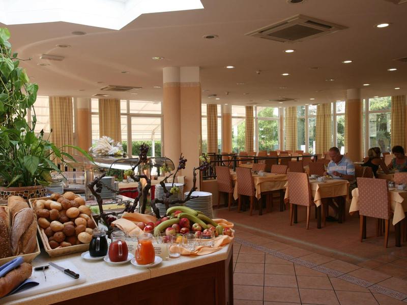 Großer heller Speisesaal mit vielen Tischen, Stühlen und einem Buffet mit Brot und Früchten.
