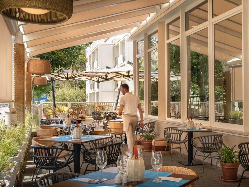 Draußen-Terrasse mit gedeckten Tischen, Stühlen und einer Person vor großen Fenstern an sonnigem Tag.