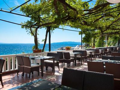 Terrasse mit Tischen und Stühlen, überdacht von Weinreben mit Blick auf das Meer bei klarem Himmel.