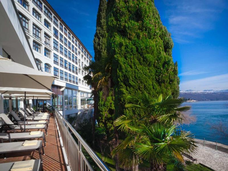 View of a hotel terrace with seating, large trees, and sea in the background.