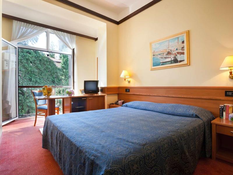 Small hotel room with double bed, desk, TV, and balcony overlooking garden.