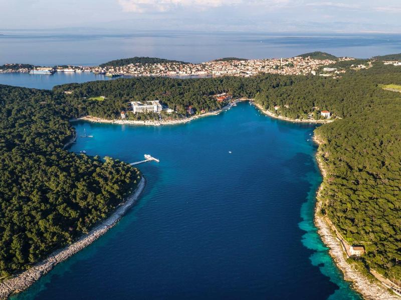 Aerial view of a forested coastal bay with clear blue water and white buildings along the shore.