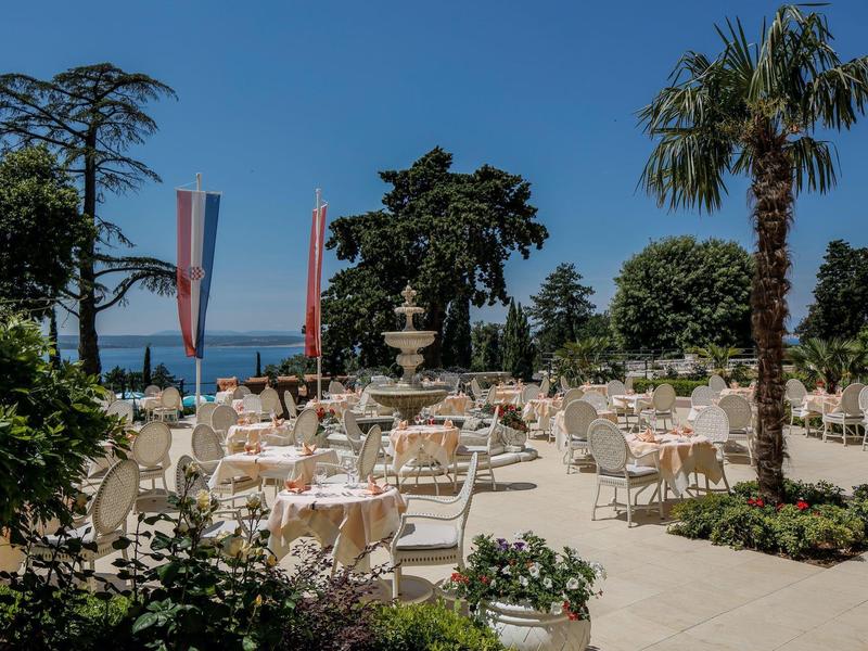 Large terrace with set tables and chairs, palm trees, and a sea view.