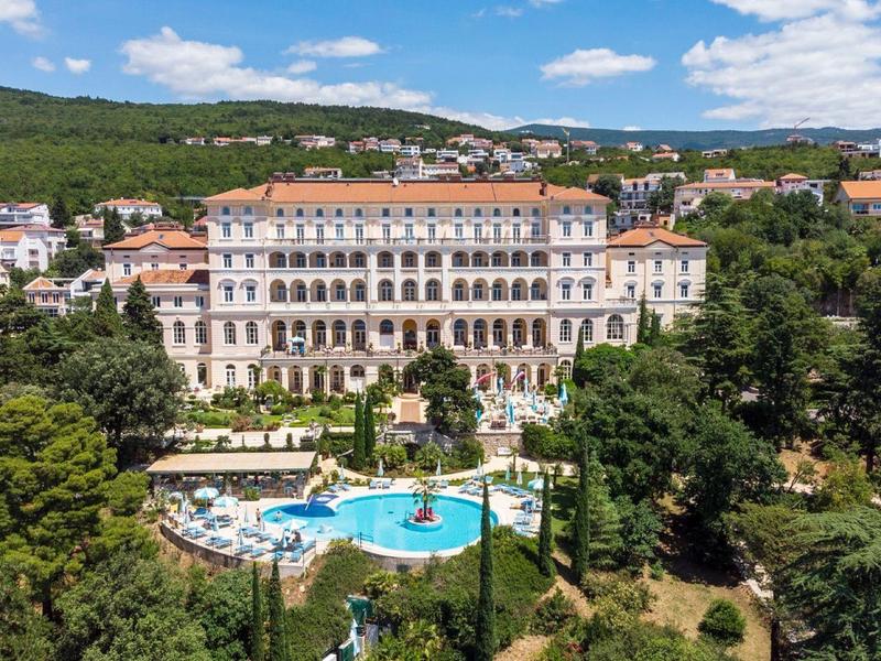 Large historic hotel with pool in green, hilly landscape under a blue sky.
