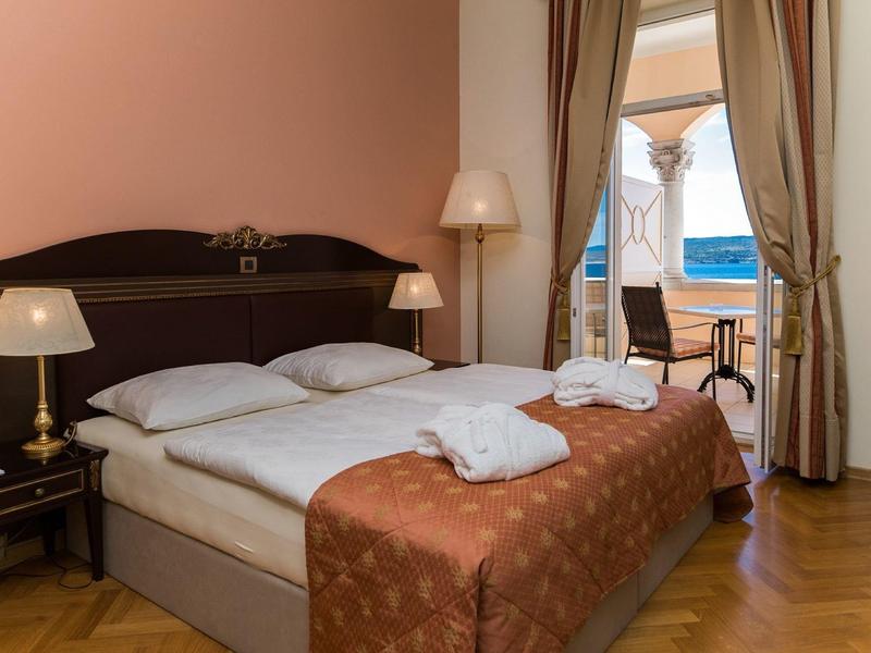 Cozy hotel room with double bed, nightstands, lamps, and sea view through open balcony door.