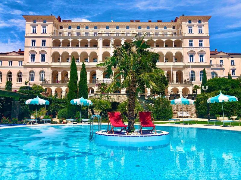 View of an elegant hotel with pool, sun umbrellas, and palm trees in the foreground
