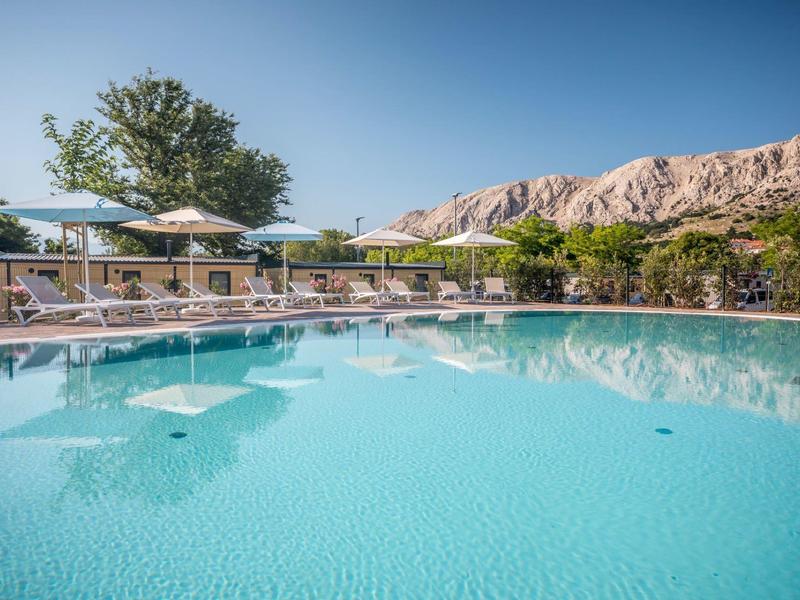 Grande piscina limpida con ombrelloni davanti a montagne sotto cielo azzurro.