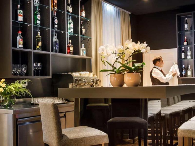 Modern bar with wooden furniture, plants, and a bartender behind the counter.