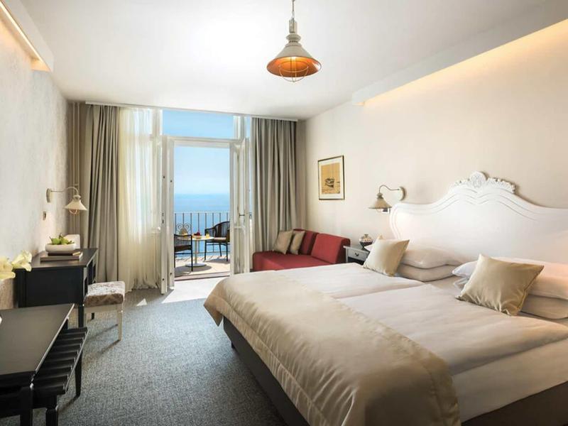 Bright hotel room with double bed, desk, sofa, and sea view through balcony windows.