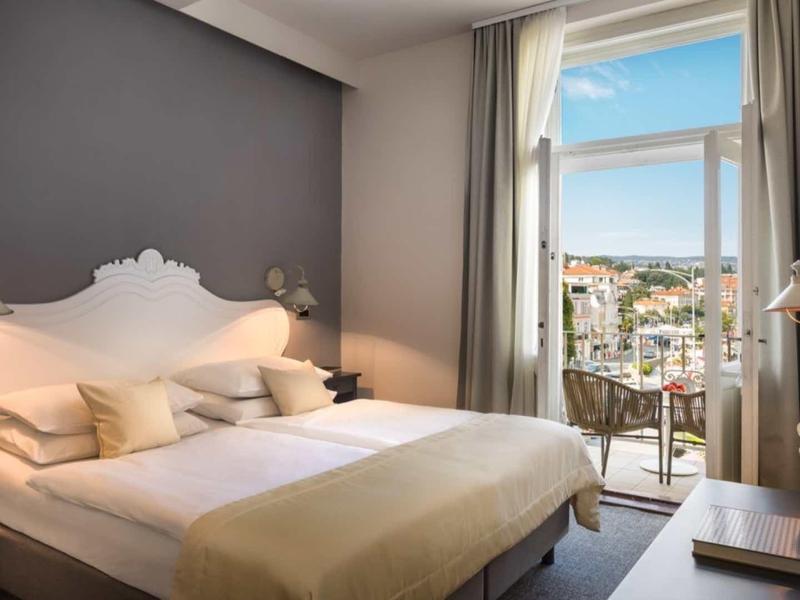 Bright hotel room with double bed, table, chair, and view of city and sea.