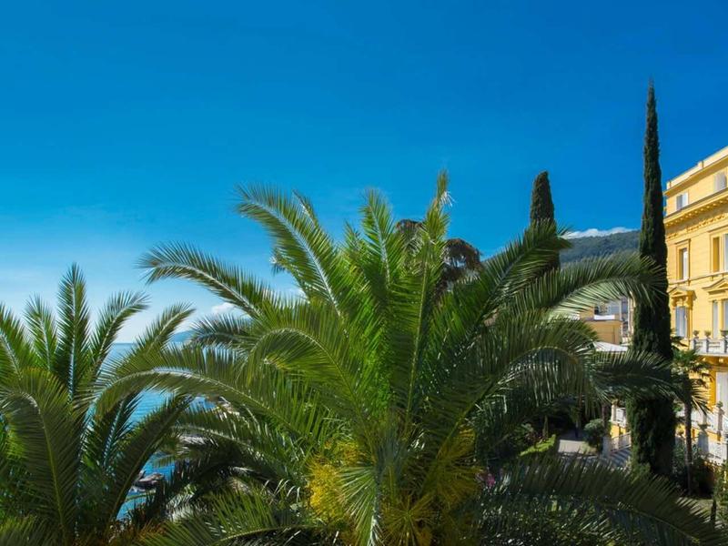 Sunlit promenade with palm trees in front of an elegant yellow building under clear sky.