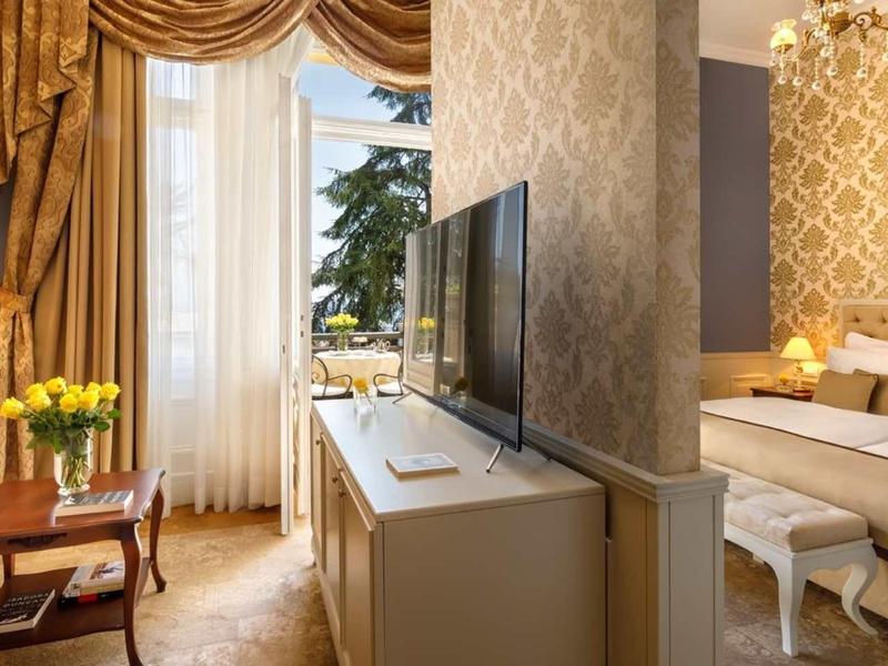 Hotel room with elegant curtain, sofa, table with yellow flowers, and bedroom with double bed and TV.