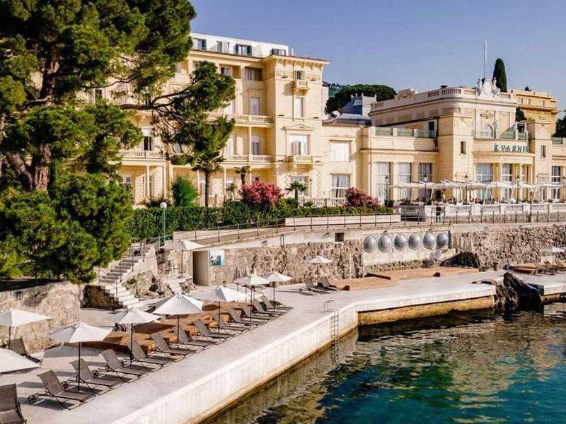 Luxury hotel by the water with terraces, sun loungers, and umbrellas under a clear sky.