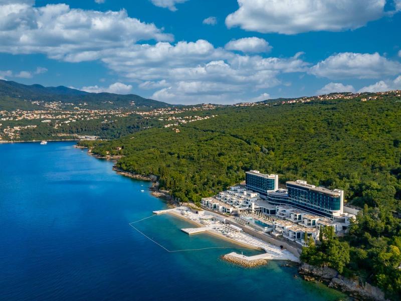 Hotel on the coast with forest, beach, and blue sea under a cloudy sky.
