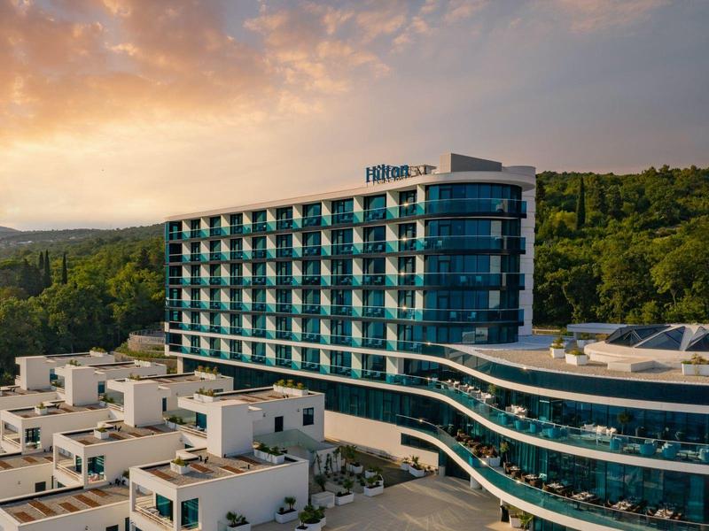 Modern hotel building with glass facade and terraces, surrounded by green hills at sunset.
