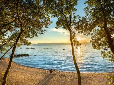 Blick durch Bäume auf einen sonnigen Strand mit zwei sitzenden Personen und ruhigem Meer.