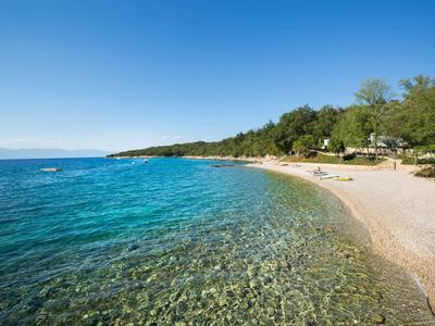 Klarer blauer Himmel über einem ruhigen Strand mit transparentem Wasser und grün bewaldetem Ufer.