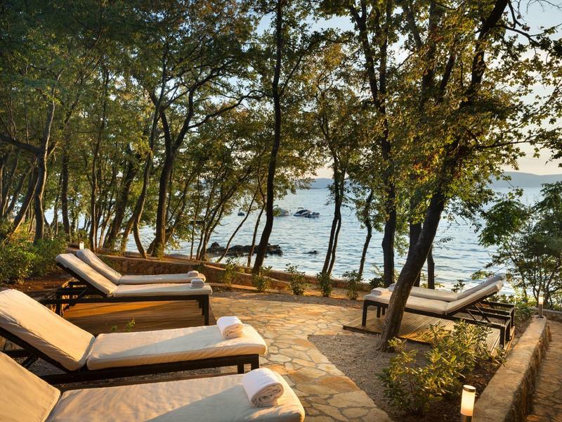 Sonnenliegen auf gepflasterter Terrasse mit Blick auf einen See und umgeben von Bäumen.