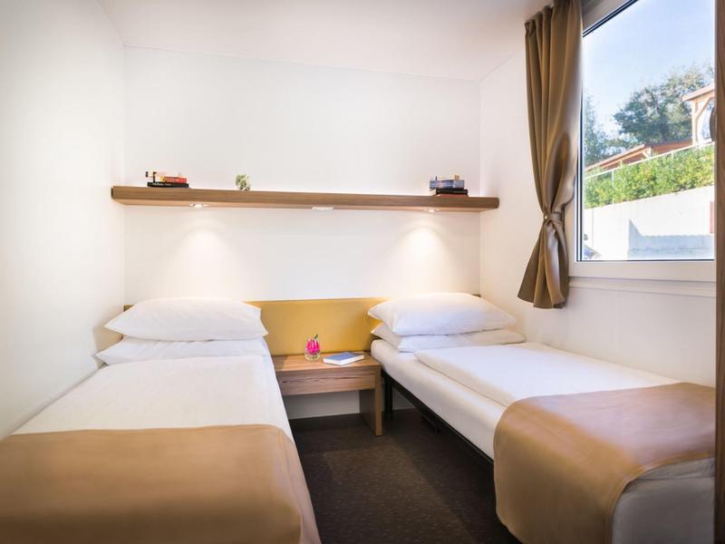 Kleines Hotelzimmer mit zwei Einzelbetten, Nachttisch und Fenster mit Blick ins Freie.