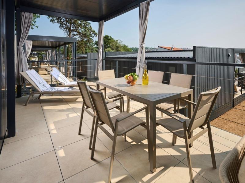 Terrasse mit Esstisch, vier Stühlen und Sonnenliegen mit Ausblick auf Bäume und Gebäude.