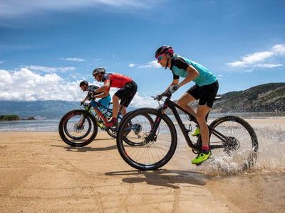 Two mountain bikers ride along the beach, one in the water, on a sunny day.