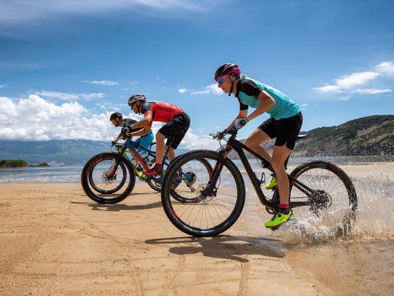 Two cyclists riding on a sandy path along a lakeshore under a clear sky.