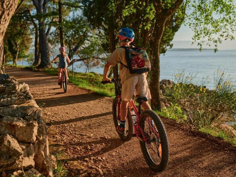 Two cyclists ride on a shaded path along a lake with trees and rocks